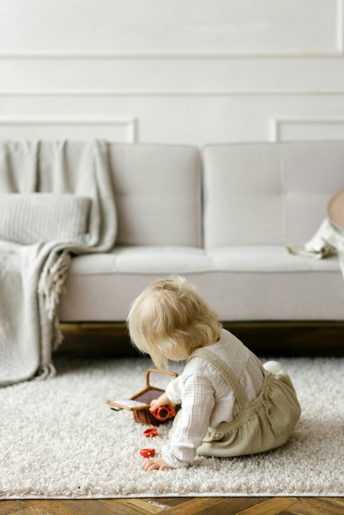 A blonde child sits on a carpet playing with toys in a cozy, neutral-toned living room.