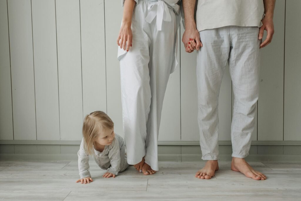 A toddler crawls on the floor while parents stand holding hands in a cozy home setting wearing casual clothes.