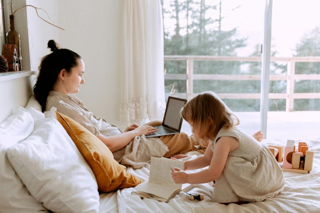 Side view of woman browsing laptop and working while spending time with daughter at home drawing in notebook